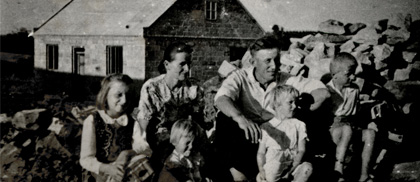 Rodzice z czwórką małych dzieci siedzą przed domem, obok góra cegieł. Archiwalne, czarno-białe zdjęcie.