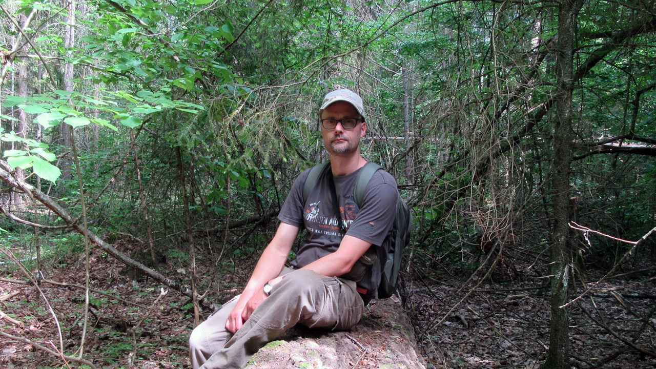 Prof. Dries Kuijper ubrany w bojówki i koszulkę siedzi na pniu powalonego drzewa
