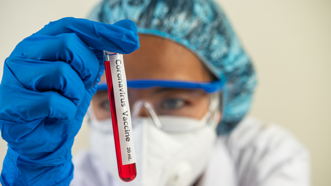 Badaczka w stroju ochronnym trzyma probówkę z napisem „szczepionka, koronawirus”